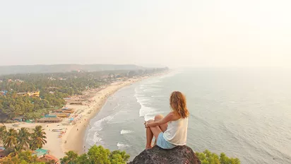 junge Frau mit blonden Haaren sitzend auf einem Felsen über dem Strand von Arambol in Goa, Indien
