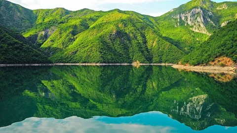 Die Landschaft um den Komansee spiegelt sich im Wasser